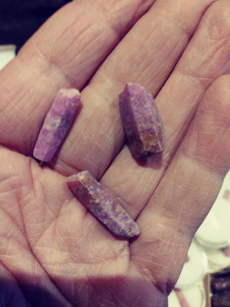 May include: Three pink and purple crystal points, with a rough, natural texture, are held in a hand.