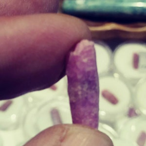 May include: A close-up of a pink and white crystal, held in a hand. The crystal is long and thin, with a smooth, polished surface.