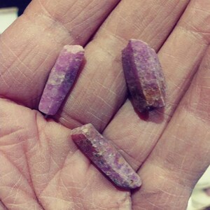 May include: Three purple and pink crystal points, also known as amethyst, are held in a hand. The crystals are long and slender with a pointed tip.