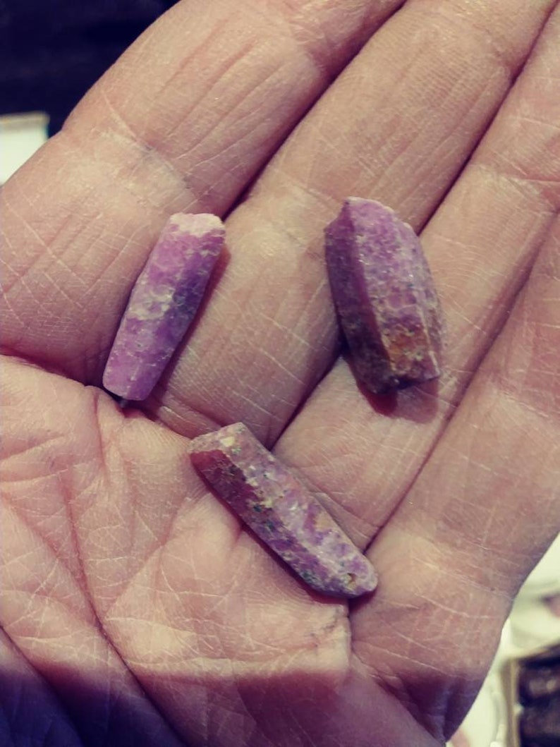 May include: Three purple and pink crystal points, also known as amethyst, are held in a hand. The crystals are long and slender with a pointed tip.