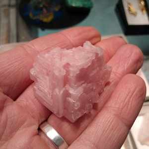 May include: A light pink, crystalline mineral specimen held in a hand. The mineral has a blocky, layered structure with a slightly translucent appearance. The background includes other display items.