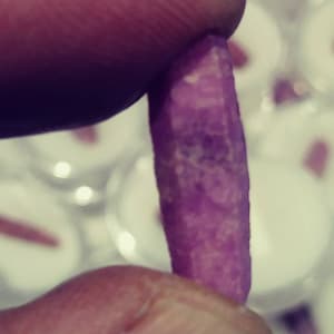 May include: A close-up of a purple gemstone, held between two fingers. The gemstone is long and thin, with a slightly irregular shape. It has a smooth, polished surface and a deep, rich purple color.