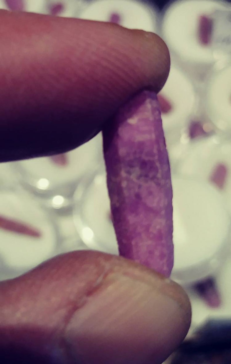 May include: A close-up of a purple gemstone, held between two fingers. The gemstone is long and thin, with a slightly irregular shape. It has a smooth, polished surface and a deep, rich purple color.