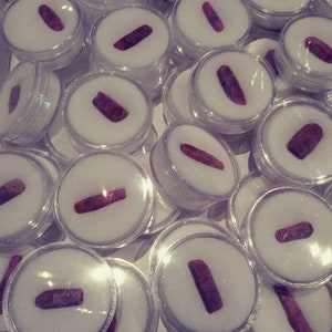 May include: A collection of small, clear plastic containers, each containing a single, long, dark red gemstone. The containers are arranged in a random pattern.