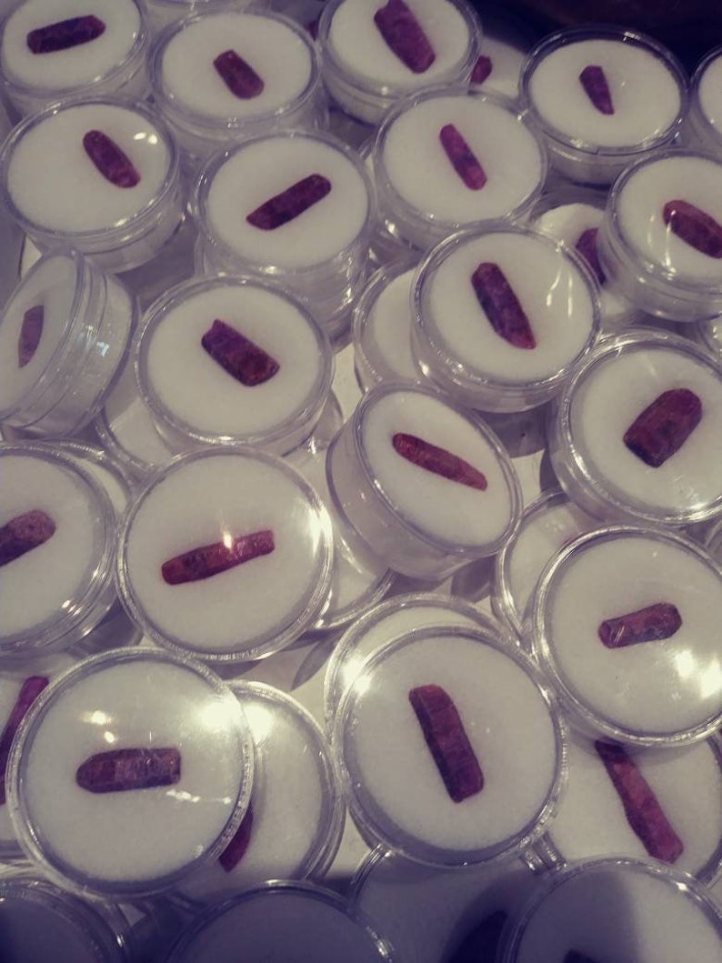 May include: A collection of small, clear plastic containers, each containing a single, long, dark red gemstone. The containers are arranged in a random pattern.