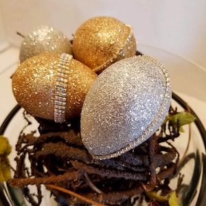 May include: Four decorative eggs in a nest. Two eggs are gold glitter, one is silver glitter, and one is gold glitter with rhinestones.