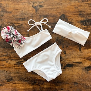 Può includere: Un set di costumi da bagno a due pezzi bianchi con un accento floreale e una fascia abbinata. Il costume da bagno presenta un dettaglio floreale a balze nei toni del rosa, bianco e nero. Il set include un top, slip e una fascia annodata.