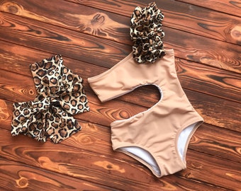 May include: A tan one-piece swimsuit with a cut-out design and a matching leopard print bow and hair accessory. The swimsuit has a white interior lining. The accessories feature a brown and black animal print.