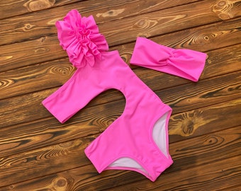 May include: A bright pink swimsuit set, including a one-shoulder swimsuit with a large floral accent and a matching headband. The swimsuit has a cutout design and white lining. The set is displayed on a wooden surface.