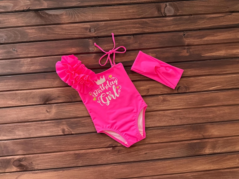 May include: A bright pink one-piece swimsuit featuring a single ruffled shoulder strap with a tie closure and a matching headband. The front of the swimsuit displays the text "Birthday Girl" in white script with a small crown and heart icons.