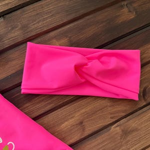 May include: A bright neon pink fabric headband featuring a central twisted knot design, positioned alongside a matching neon pink garment with visible yellow graphic text and thin pink straps.