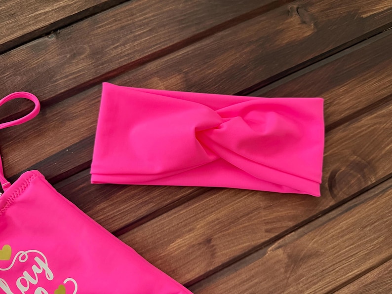 May include: A bright neon pink fabric headband featuring a central twisted knot design, positioned alongside a matching neon pink garment with visible yellow graphic text and thin pink straps.