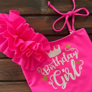May include: A neon pink one-piece swimsuit featuring a large ruffled shoulder detail, thin adjustable straps, and the white printed text "Birthday Girl" accompanied by a small crown icon and several small green heart graphics on the front.