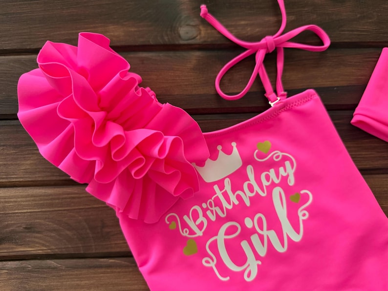 May include: A neon pink one-piece swimsuit featuring a large ruffled shoulder detail, thin adjustable straps, and the white printed text "Birthday Girl" accompanied by a small crown icon and several small green heart graphics on the front.