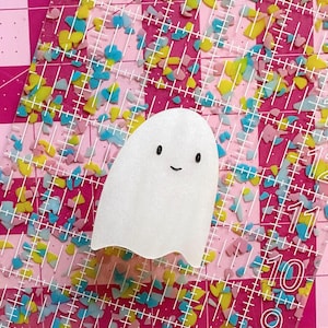 May include: A white ghost-shaped craft with a smiling face. The ghost is on a pink cutting mat with colorful confetti and white grid lines. The mat has measurements in inches.