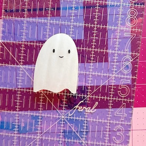 May include: A white ghost-shaped sticker with two black oval eyes and a small black curved mouth, positioned on a clear acrylic quilting ruler with white grid markings and numbers, showing a height of approximately 2 inches.