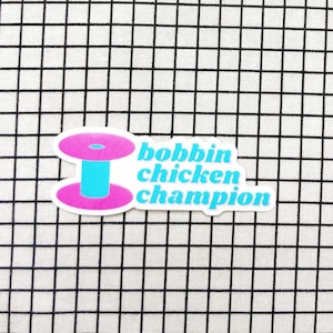 May include: A die-cut sticker featuring a bright pink and light blue sewing bobbin graphic next to the light blue text that reads bobbin chicken champion. The sticker has a white border and is displayed against a white fabric with a black grid.