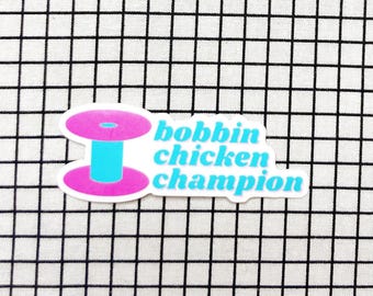 Może przedstawiać: Wykrawana naklejka z grafiką jaskraworóżowej i jasnoniebieskiej szpulki do nici obok jasnoniebieskiego napisu bobbin chicken champion. Naklejka ma białą obwódkę i jest umieszczona na białej tkaninie w czarną kratkę.