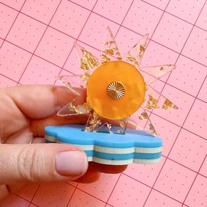 May include: A whimsical, layered acrylic sunburst design with gold leaf accents. The sun is centered on an orange circle with a gold center, atop a blue and white cloud-shaped base. The design is held against a pink cutting mat.
