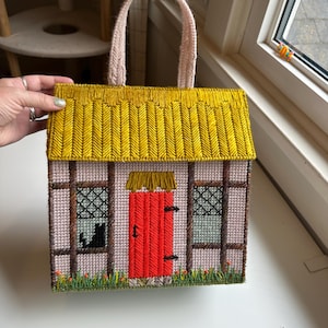 Puede incluir: Bolso tote artesanal con forma de cabaña. Cuenta con un tejado amarillo, una puerta roja y ventanas con silueta de gato negro. El asa es rosa y el bolso está hecho de un material tejido.
