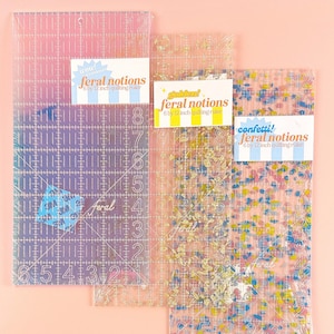 May include: Three clear quilting rulers, each 6 by 12 inches, with different designs. One ruler has a gradient of pink and purple, another has gold floral accents, and the third has colorful confetti. Each ruler has the text "feral notions" and "6 by 12 inch quilting ruler".