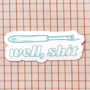 May include: A white sticker with a teal illustration of a sewing needle and the words "well, shit" in a retro font. The sticker is on a red and white checkered background.