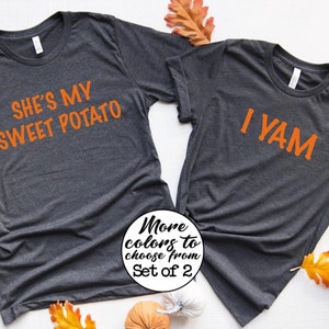 May include: Two charcoal grey t-shirts with orange text. One reads "She's my sweet potato" and the other says "I yam". The image includes autumn leaves and a small pumpkin. The text "More colours to choose from Set of 2" is also visible.