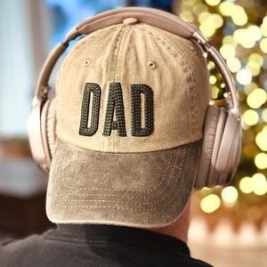 Op de afbeelding: Een kaki baseballpet met het woord "DAD" geborduurd in donkergroen. De pet wordt gedragen met over-ear koptelefoon. De persoon draagt een zwart shirt.