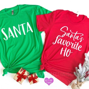 Pu&ograve; includere: Un set di due magliette a tema natalizio. Una maglietta verde dice "SANTA" in testo bianco. L'altra maglietta rossa dice "Santa's Favorite Ho" in testo bianco.