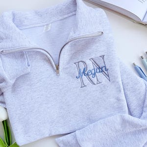 May include: A light gray heathered quarter-zip sweatshirt featuring an embroidered design on the left chest. The embroidery consists of large gray block letters RN with the name Megan stitched in blue cursive script across the center of the letters.