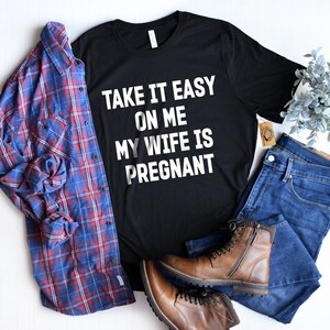 May include: Black t-shirt with white text that reads "Take it easy on me my wife is pregnant". The shirt is styled with a blue plaid shirt, blue jeans, and brown boots.