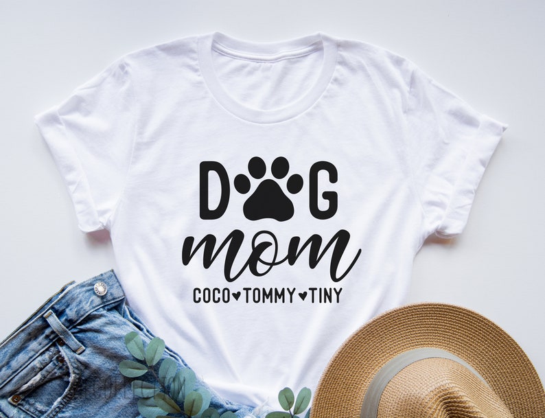 May include: White t-shirt with black text that reads "Dog Mom" with a paw print between the words. Below the text is "Coco Tommy Tiny" with a heart between each name.