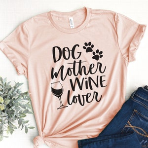 May include: A light pink t-shirt with the words "Dog Mother Wine Lover" in black script. The design includes paw prints and a wine glass. The shirt is paired with blue jeans.