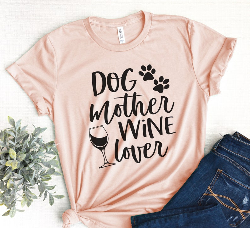 May include: A light pink t-shirt with the words "Dog Mother Wine Lover" in black script. The design includes paw prints and a wine glass. The shirt is paired with blue jeans.