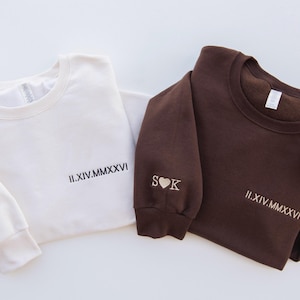 May include: Two crewneck sweatshirts, one cream and one brown, are displayed. The cream sweatshirt has the Roman numeral date "II.XIV.MMXXVI" and the initials "S K" with a heart. The brown sweatshirt also has the date and initials.