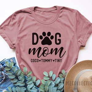 May include: A dusty rose t-shirt with the text "Dog Mom" in black lettering. A paw print is between the "D" and the "G". Below the text is "Coco Tommy Tiny" with a heart between each name.