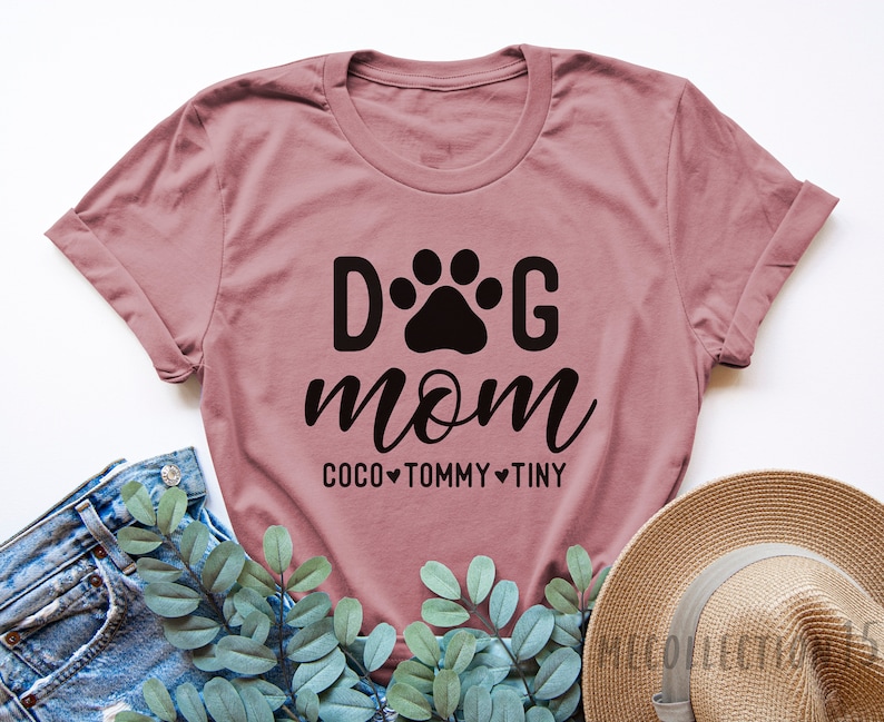 May include: A dusty rose t-shirt with the text "Dog Mom" in black lettering. A paw print is between the "D" and the "G". Below the text is "Coco Tommy Tiny" with a heart between each name.