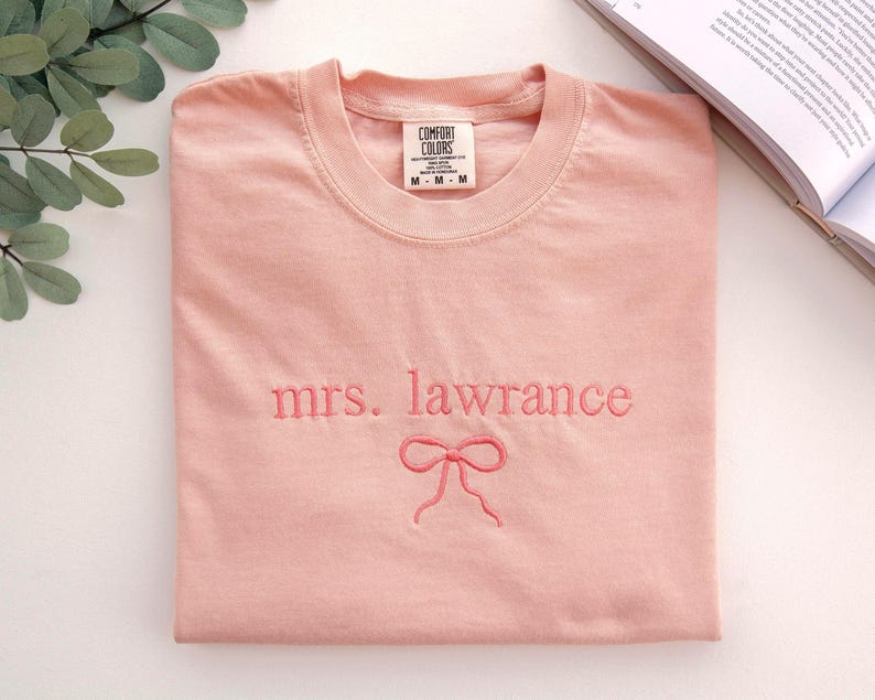 Puede incluir: Camiseta rosa claro con bordado "mrs. lawrance" y un lazo. La camiseta est&aacute; doblada, con una etiqueta visible en el cuello. La tela parece suave y c&oacute;moda.