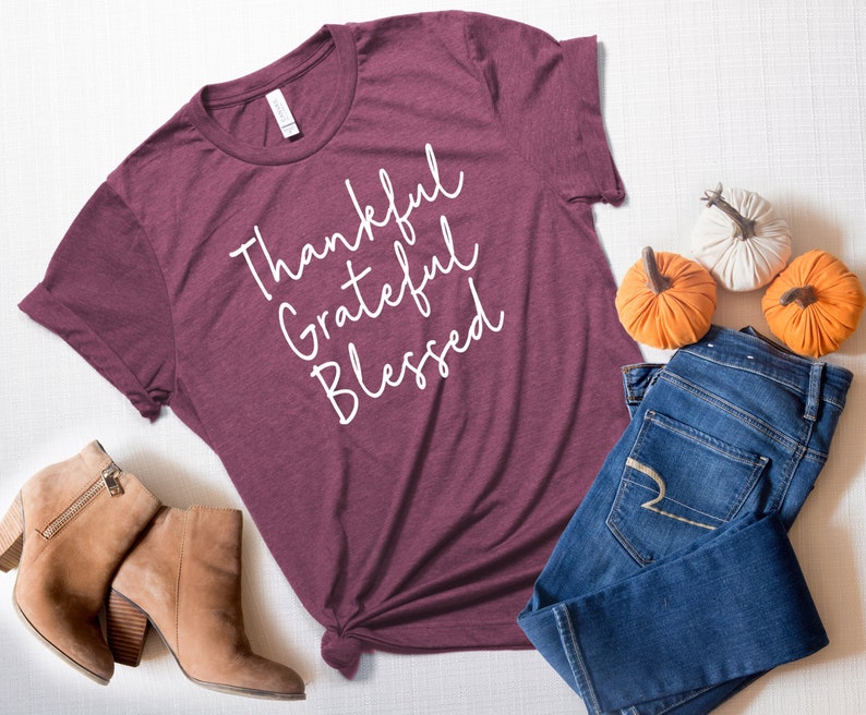 K&ouml;nnte beinhalten: Ein maroonfarbenes T-Shirt mit den Worten "Thankful Grateful Blessed" in wei&szlig;er Kursivschrift. Das Shirt liegt auf einer wei&szlig;en Oberfl&auml;che mit einem Paar braunen Stiefeletten, einer blauen Jeans und drei kleinen K&uuml;rbissen.