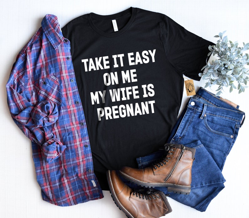 May include: Black t-shirt with white text that reads "Take it easy on me my wife is pregnant". The shirt is styled with a blue plaid shirt, blue jeans, and brown boots.