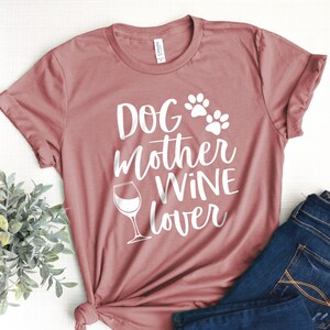 May include: Dusty rose t-shirt with the words "DOG MOTHER WINE lover" in white script, along with paw prints and a wine glass graphic. The shirt is folded, with a pair of blue jeans visible in the background.