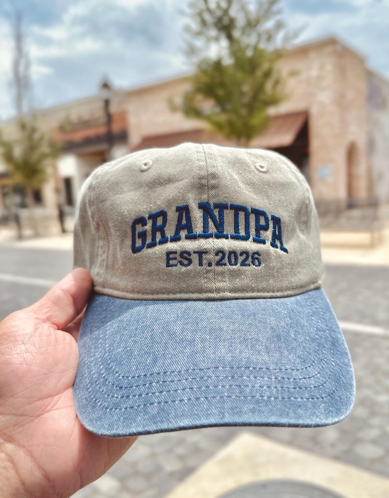 K&ouml;nnte beinhalten: Beige und blaue Baseballkappe mit dem Wort "GRANDPA" in Blau gestickt, zusammen mit "EST. 2026". Die Kappe hat einen gebogenen Schirm und ein klassisches sechsteiliges Design. Ein l&auml;ssiges Accessoire.