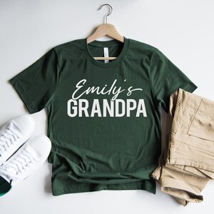 以下が含まれることがあります： 白い文字で「Emily's GRANDPA」と書かれた濃い緑色のTシャツです。