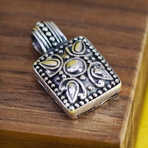 May include: A silver pendant with an ornate, rectangular design. The pendant features a series of raised, circular and teardrop-shaped elements, surrounded by small, beaded details. The bail is a simple loop with a beaded texture. The pendant is likely for a necklace.