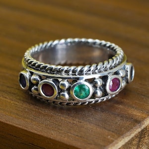 May include: A silver ring with a twisted rope design and a row of alternating gemstones. The ring features oval-cut gemstones in purple, red, and green. The ring is set on a wooden surface.