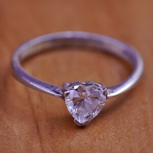 May include: A silver ring with a heart-shaped, clear gemstone. The ring's band is a simple, polished silver. The gemstone is set in a four-prong setting, and the ring is displayed on a wooden surface.