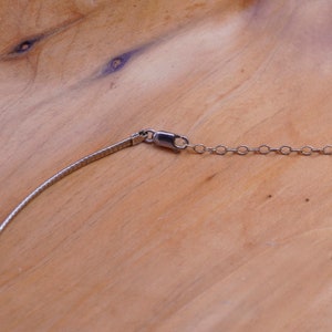 May include: A silver-toned necklace with a textured, flat chain and a small chain of circular links. The necklace has a lobster clasp closure. The background is a light brown wooden surface.