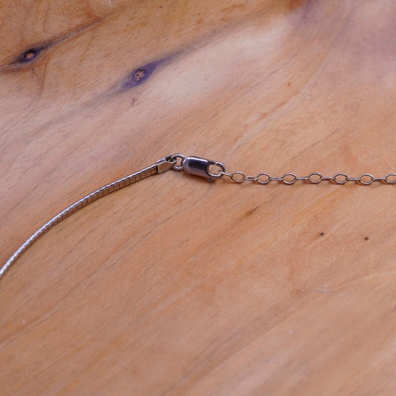 May include: A silver-toned necklace with a textured, flat chain and a small chain of circular links. The necklace has a lobster clasp closure. The background is a light brown wooden surface.