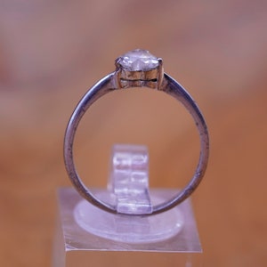 May include: A silver ring with a pear-shaped, clear gemstone. The ring is set on a clear acrylic stand against a blurred, warm-toned background. The band of the ring is thin and appears to be made of a silver-colored metal.