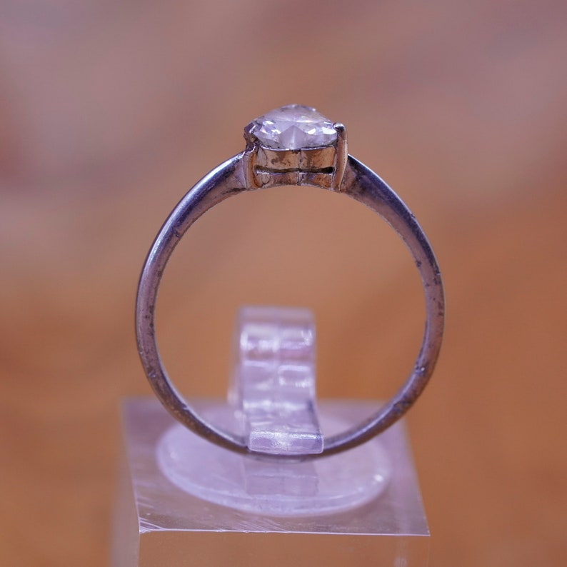 May include: A silver ring with a pear-shaped, clear gemstone. The ring is set on a clear acrylic stand against a blurred, warm-toned background. The band of the ring is thin and appears to be made of a silver-colored metal.